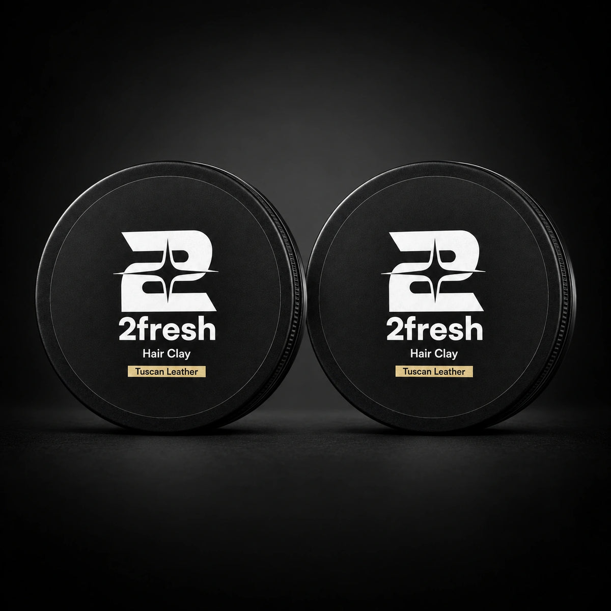 Two closed 2Fresh Hair Clay tins in Tuscan Leather on a dark studio background